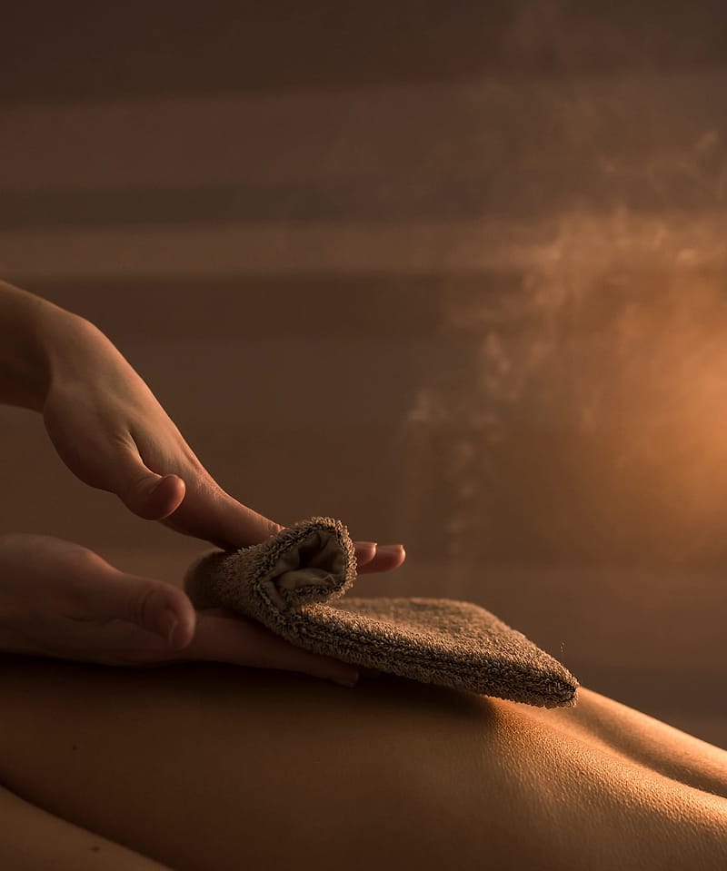 A therapist gently wipes a warm towel mitt over a guest’s back in the steam